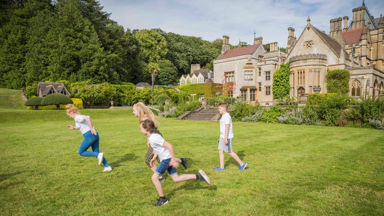 Four children running around on grass outside the house at Tyntesfield in the summer, with the house in the background